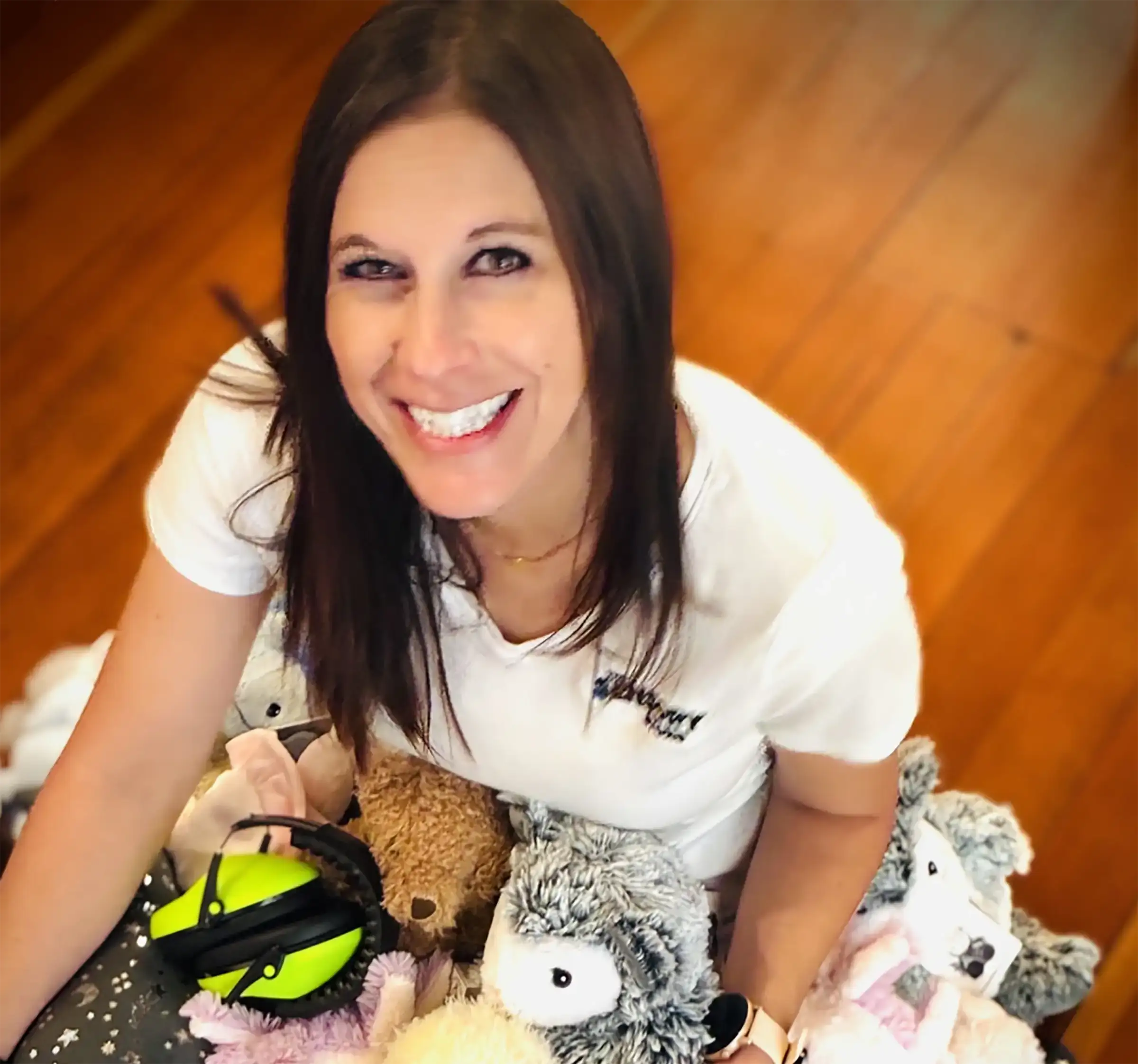Samantha sitting on the floor with a bunch of Warmies stuffies and other products on her lap, smiling at the camera