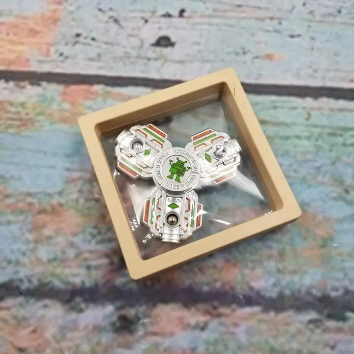 silver metal 3-pronged spinner with green robot at center in clear box with beige frame