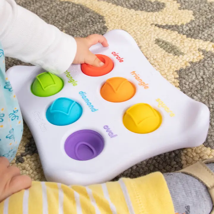 white rectangular toy with 6 different bright colored buttons with shapes on them and a toddler pushing some of the buttons in