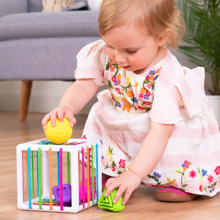 toddler pushing a yellow sphere through the multicolored elastic bands on a White cube