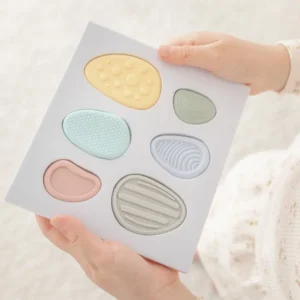 two hands hold a package card with 6 pastel colored silicone worry stones with different shapes, sizes and textures nestled inside