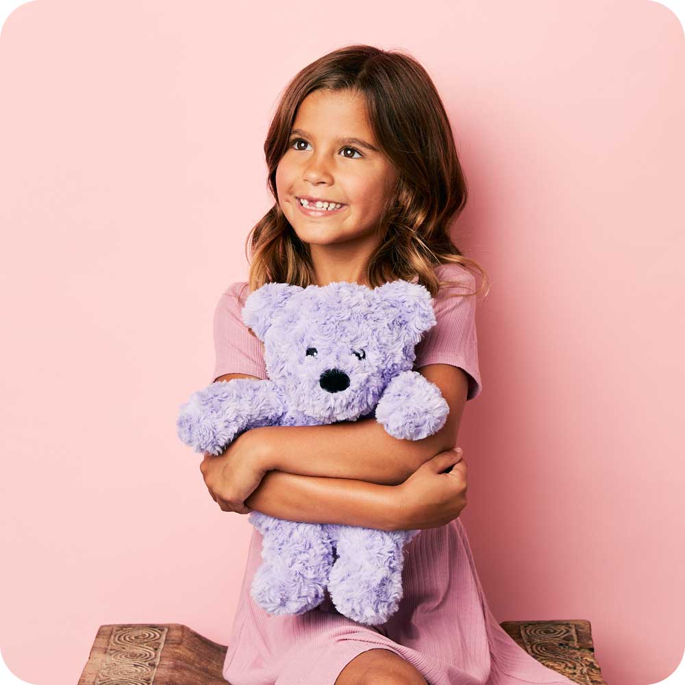 girl holding lavender colored teddy bear with short curly fur against a pink wall